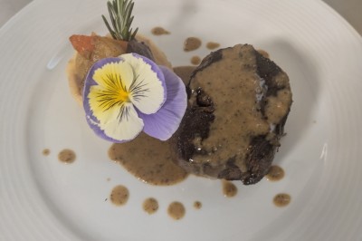 Assiette avec un morceau de viande nappé de sauce, purée, garniture de légumes et fleur comestible en décoration.