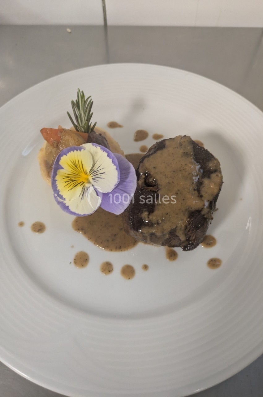 Assiette avec un morceau de viande nappé de sauce, purée, garniture de légumes et fleur comestible en décoration.
