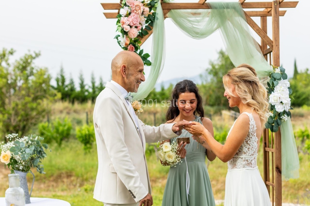 Un couple échange des alliances sous une arche décorée de fleurs, accompagné d'une femme tenant un bouquet.