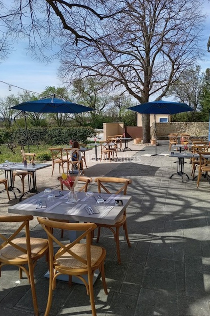 Terrasse ensoleillée avec tables et chaises en bois, parasols bleus et vue sur un paysage verdoyant.