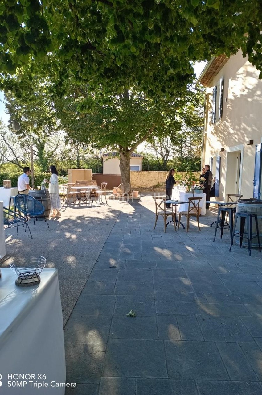 Terrasse ensoleillée avec des tables et chaises disposées sous un grand arbre, quelques personnes en arrière-plan.