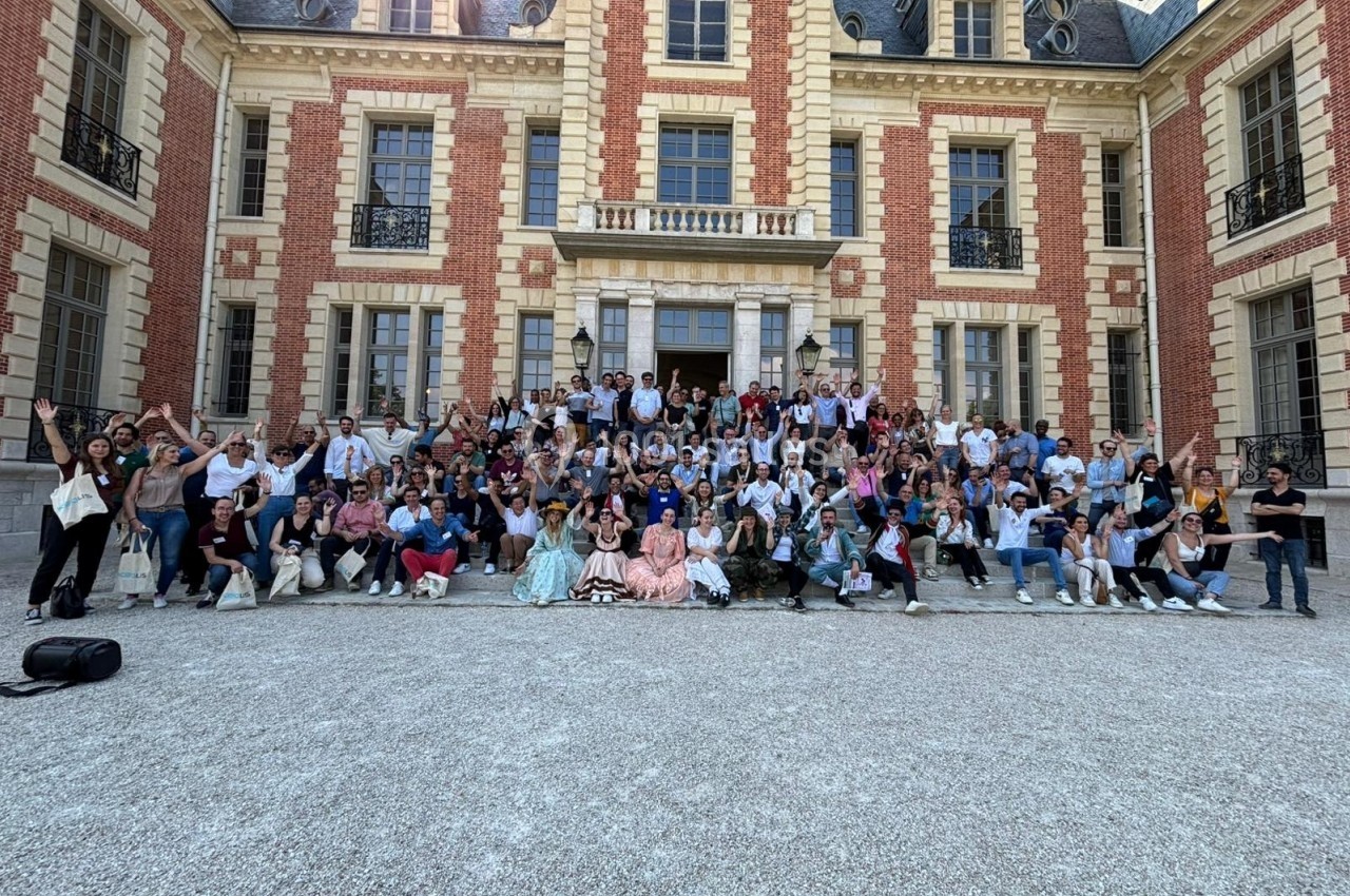Groupe de personnes posant devant un grand bâtiment en briques rouges et pierres claires, par une journée ensoleillée.