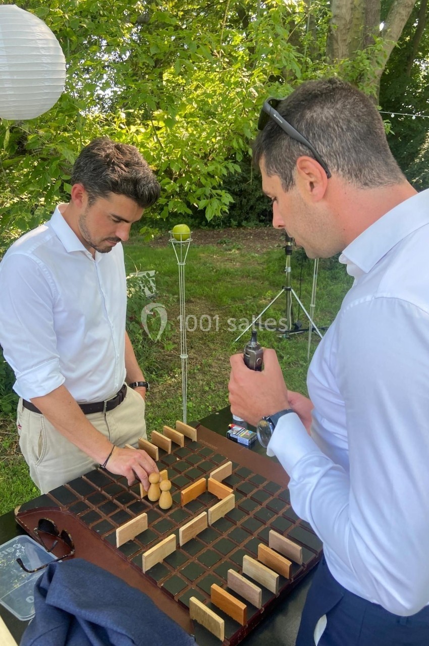 Deux hommes jouent à un jeu de société en bois sur une table en extérieur, entourés de verdure.
