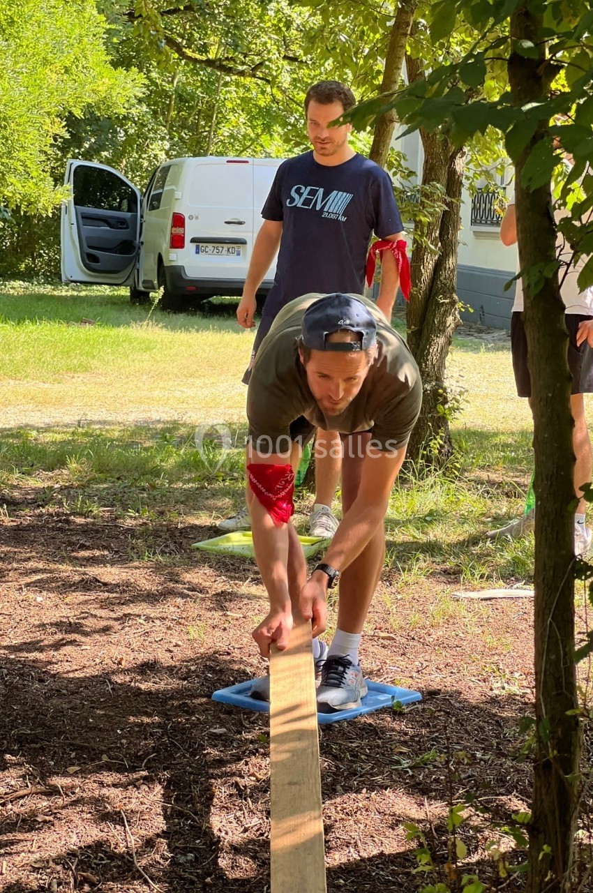 Un homme en équilibre sur une planche en bois posée au sol, entouré de participants dans un espace extérieur boisé.
