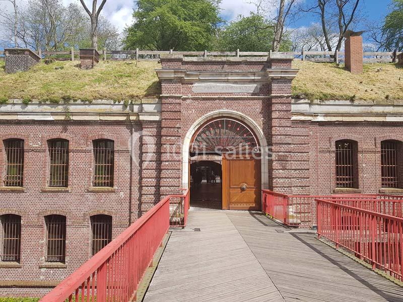 Entrée principale d'un bâtiment en briques rouges avec une arche, vue depuis une passerelle en bois et métal rouge.