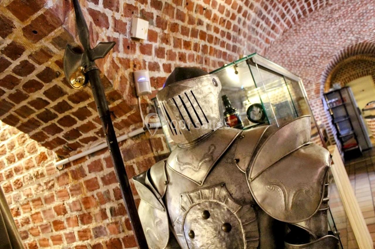 Armure médiévale en métal exposée dans une pièce voûtée en briques avec une vitrine en arrière-plan.