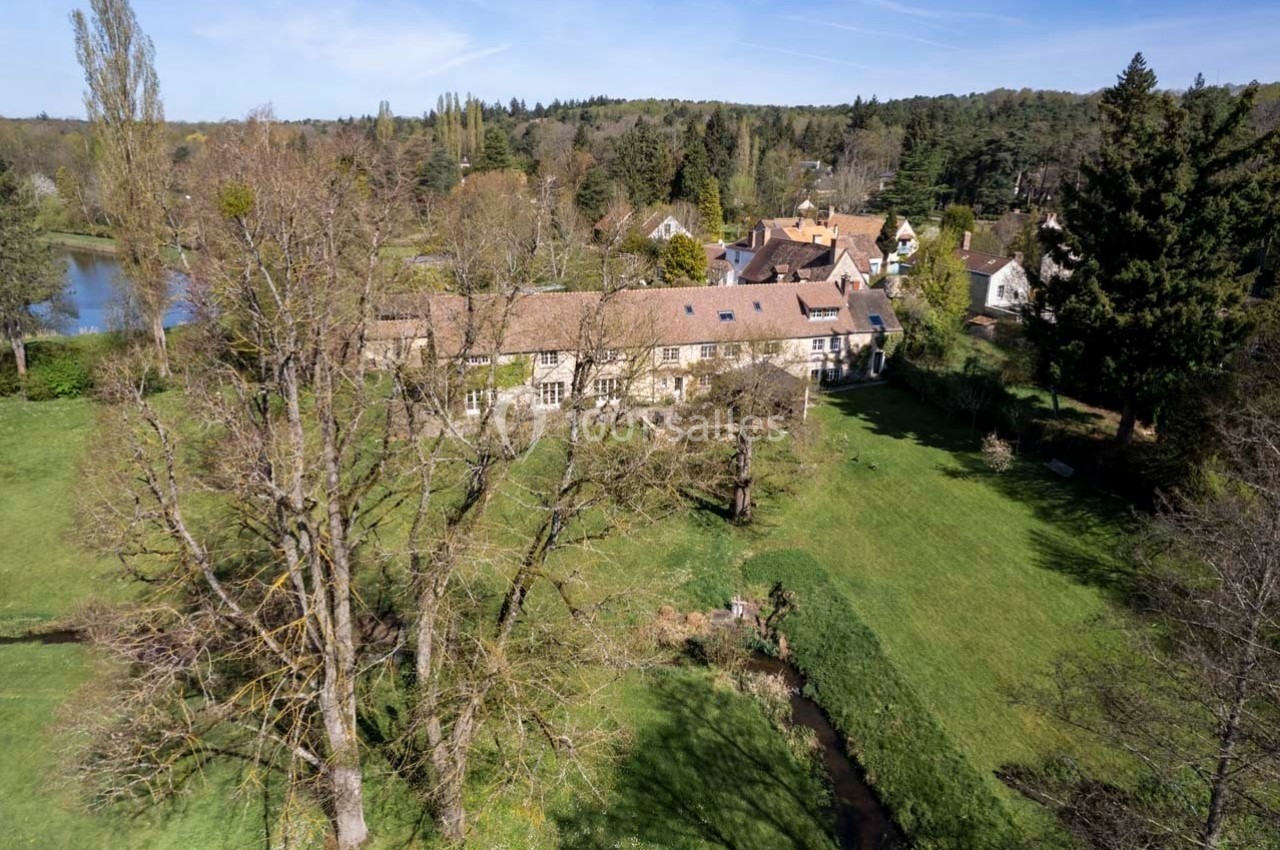 Vue aérienne d'une maison en pierre entourée d'arbres, d'une pelouse verdoyante et d'un ruisseau, avec un étang à proximité.