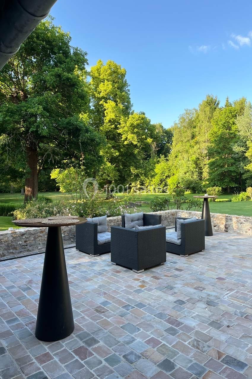 Mobilier de jardin en rotin noir avec coussins gris sur une terrasse en pierre, entourée d'un parc verdoyant.