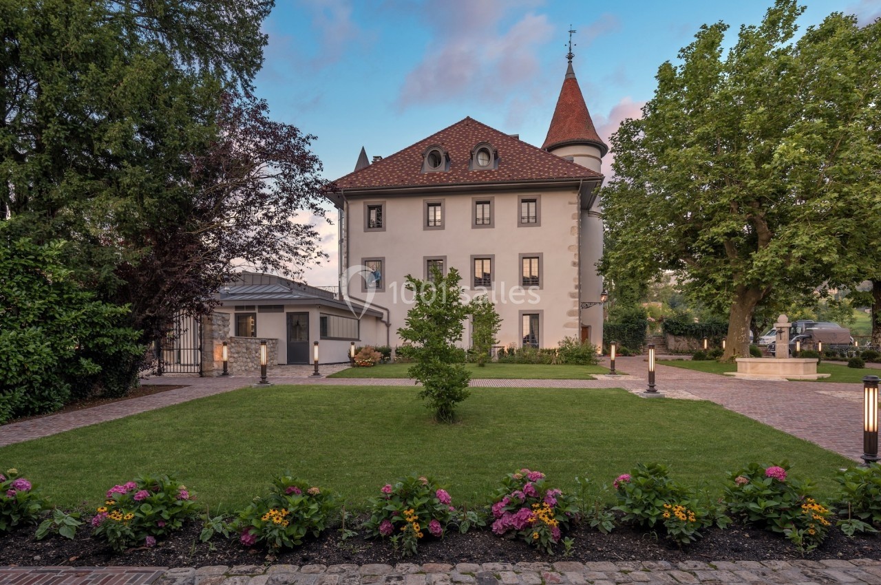 Façade d'un bâtiment historique avec toit en tuiles rouges, entouré d'un jardin fleuri et d'arbres.