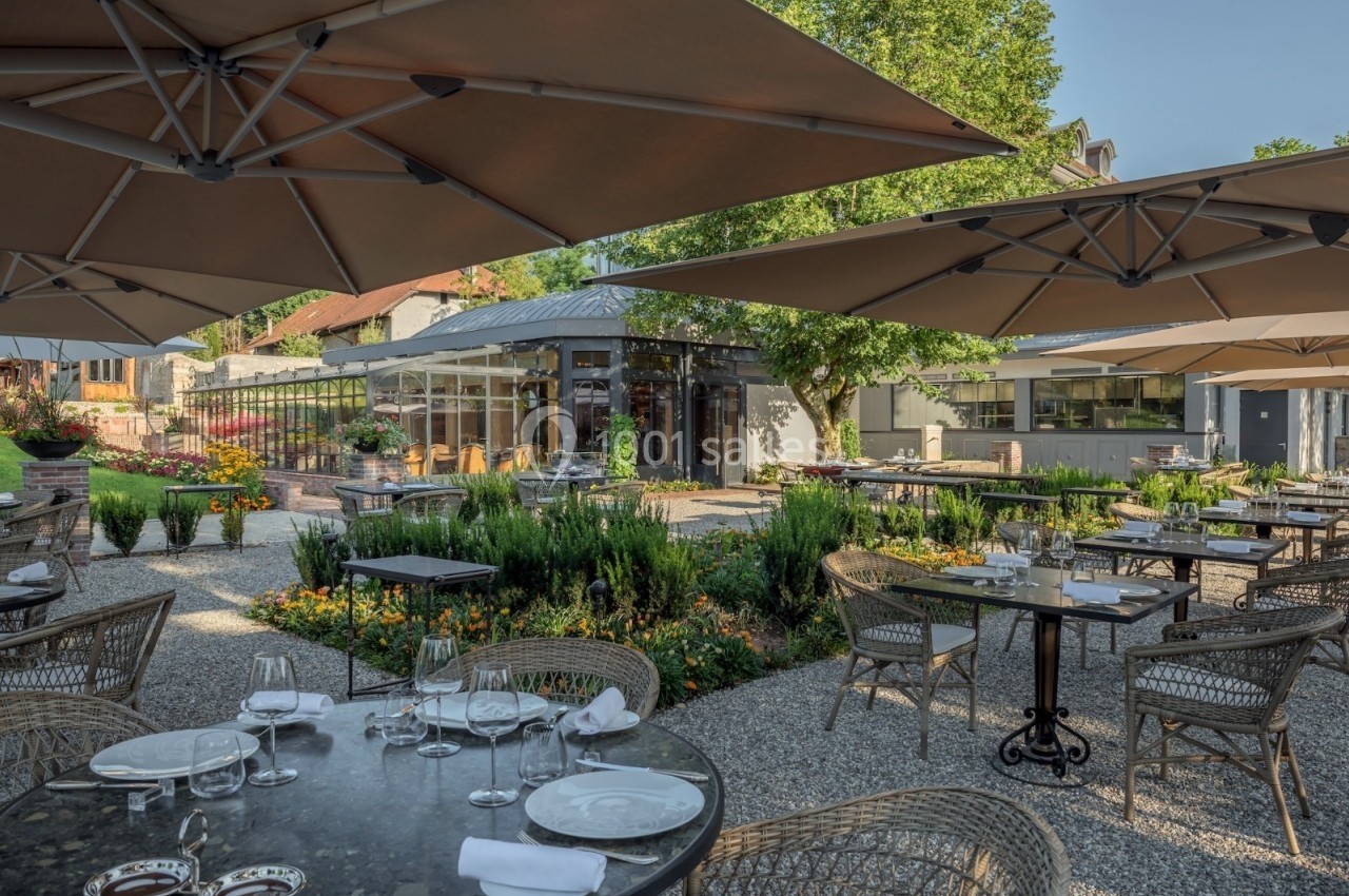 Terrasse d'un restaurant avec tables dressées, chaises en osier, parasols et jardin fleuri sous un ciel dégagé.