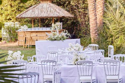 Coussin brodé ’Villa Alexandra’ au premier plan, banc en bois flou en arrière-plan dans un jardin ensoleillé.