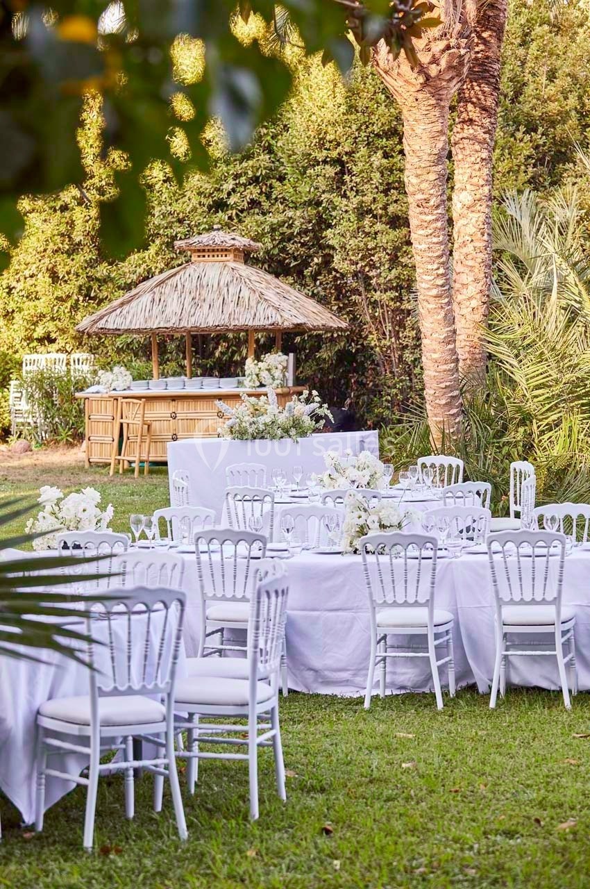 Tables blanches décorées pour un événement en plein air, entourées de verdure avec un bar en bambou en arrière-plan.