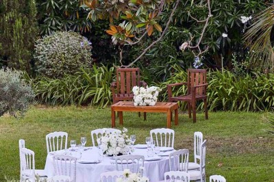 Coussin brodé ’Villa Alexandra’ au premier plan, banc en bois flou en arrière-plan dans un jardin ensoleillé.