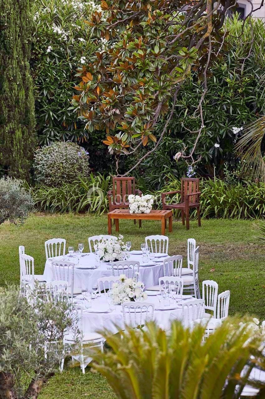 Tables blanches décorées pour un événement en plein air, entourées de verdure et d'arbres dans un jardin.