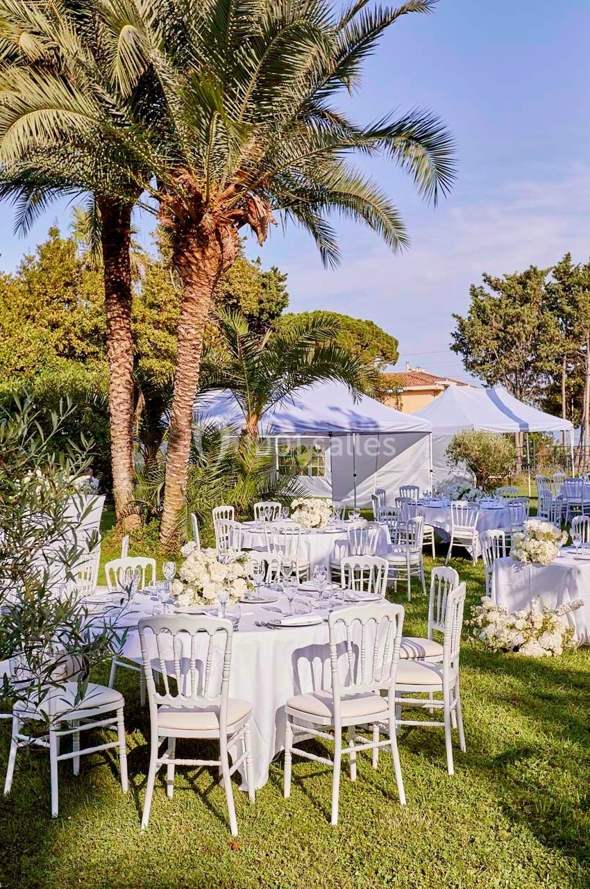 Tables décorées de fleurs blanches disposées sur une pelouse, entourées de palmiers et de tentes blanches.