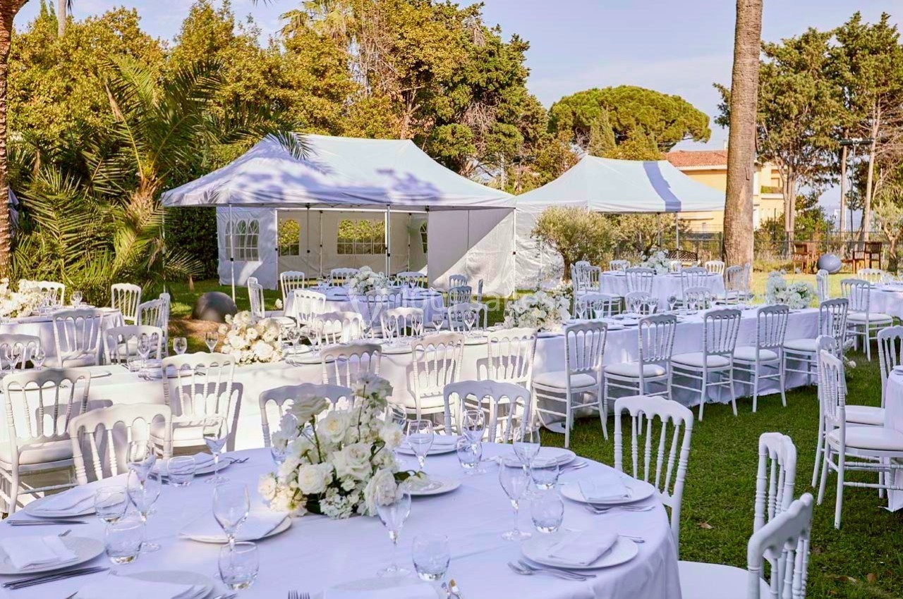 Tables dressées avec nappes blanches et chaises disposées dans un jardin, avec des tentes blanches en arrière-plan.
