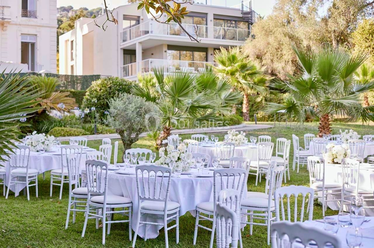 Tables rondes décorées de nappes blanches et chaises alignées dans un jardin verdoyant près d'un bâtiment moderne.