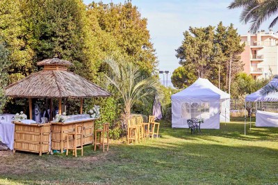 Coussin brodé ’Villa Alexandra’ au premier plan, banc en bois flou en arrière-plan dans un jardin ensoleillé.