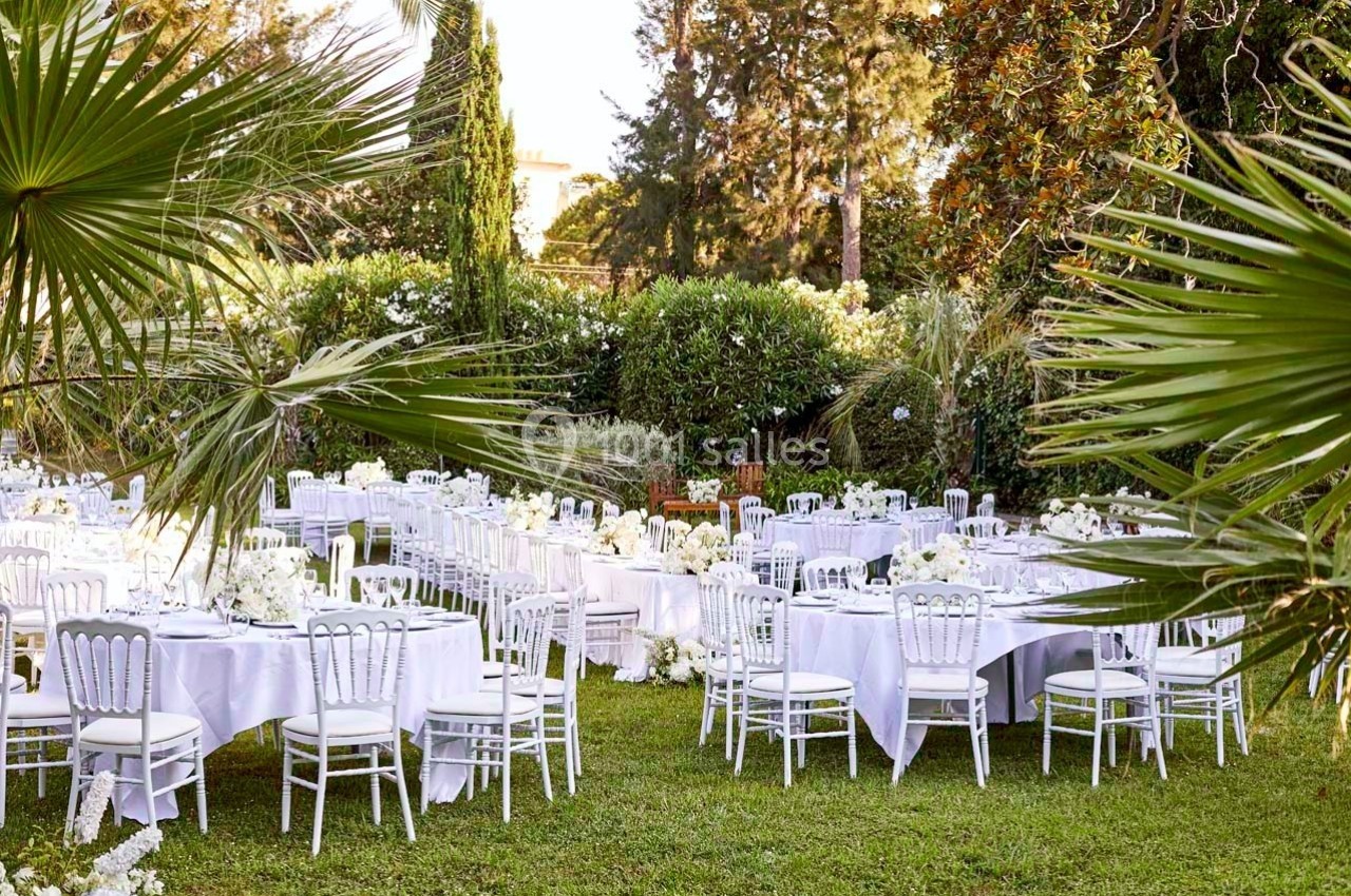 Tables rondes décorées de nappes blanches et chaises disposées dans un jardin verdoyant pour un événement en plein air.