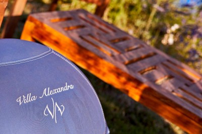 Coussin brodé ’Villa Alexandra’ au premier plan, banc en bois flou en arrière-plan dans un jardin ensoleillé.