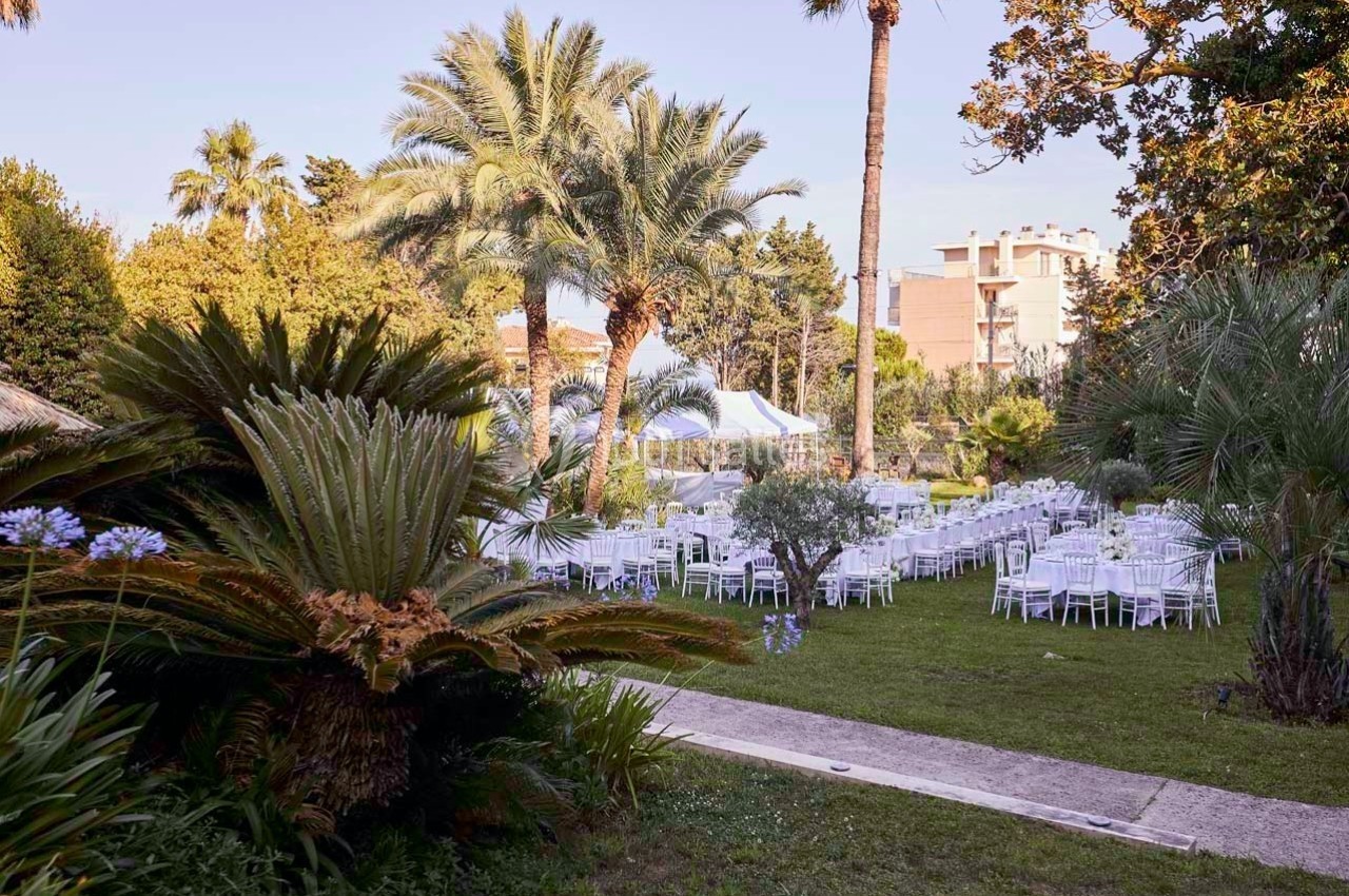 Jardin aménagé avec des tables et chaises blanches disposées pour un événement, entouré de palmiers et de végétation.