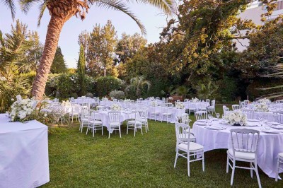 Coussin brodé ’Villa Alexandra’ au premier plan, banc en bois flou en arrière-plan dans un jardin ensoleillé.