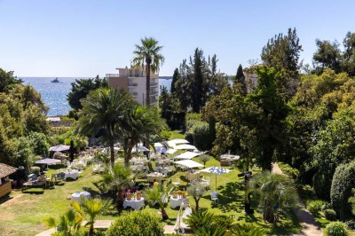 Coussin brodé ’Villa Alexandra’ au premier plan, banc en bois flou en arrière-plan dans un jardin ensoleillé.