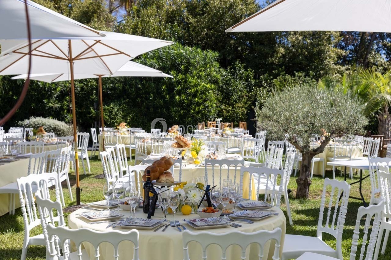 Tables dressées en extérieur avec nappes claires, chaises blanches, parasols et décorations florales sur pelouse verdoyante.