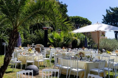 Coussin brodé ’Villa Alexandra’ au premier plan, banc en bois flou en arrière-plan dans un jardin ensoleillé.