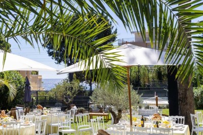 Coussin brodé ’Villa Alexandra’ au premier plan, banc en bois flou en arrière-plan dans un jardin ensoleillé.