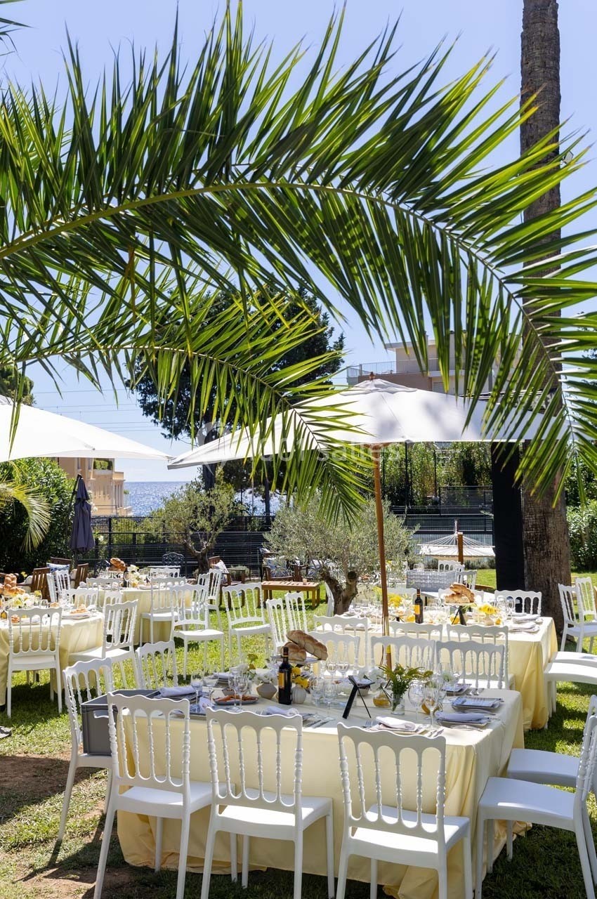 Tables dressées avec nappes jaunes et chaises blanches dans un jardin ensoleillé, entouré de palmiers et de parasols.