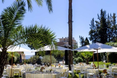 Coussin brodé ’Villa Alexandra’ au premier plan, banc en bois flou en arrière-plan dans un jardin ensoleillé.