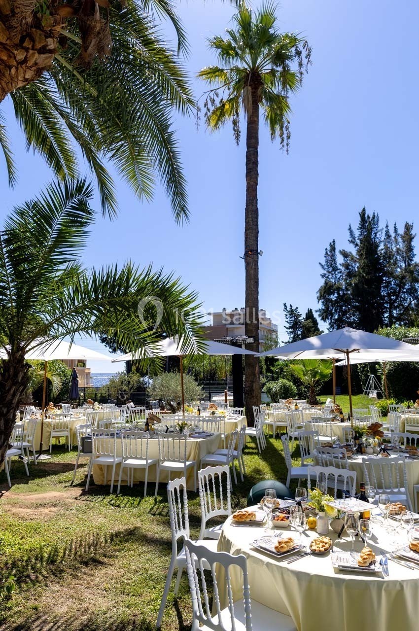 Tables dressées avec nappes blanches et chaises disposées dans un jardin ensoleillé, entouré de palmiers et de parasols.