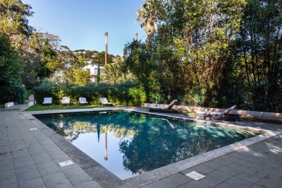 Coussin brodé ’Villa Alexandra’ au premier plan, banc en bois flou en arrière-plan dans un jardin ensoleillé.