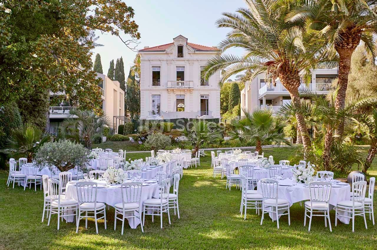 Tables dressées avec nappes blanches et chaises disposées dans un jardin devant une grande maison de style classique.