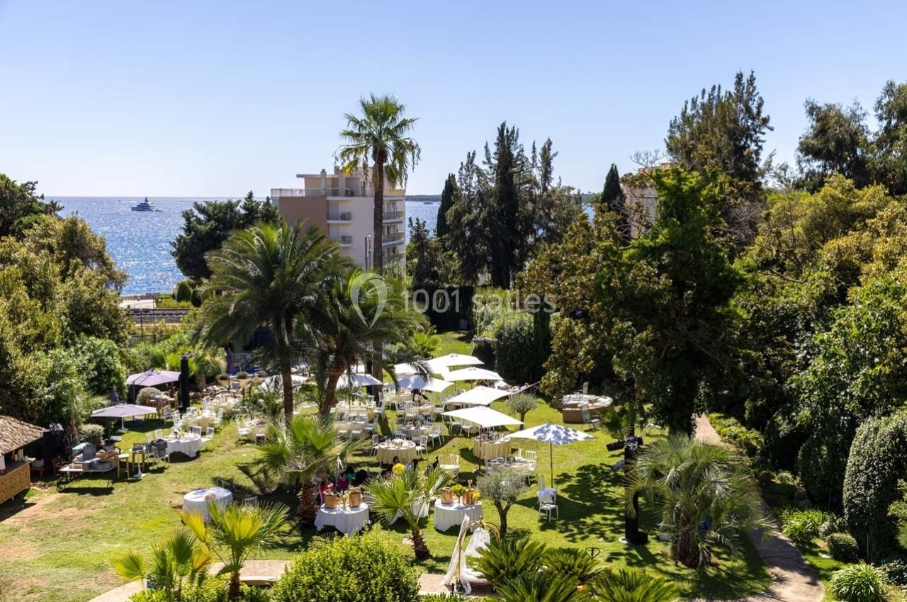 Jardin avec tables dressées pour un repas en plein air, entouré de palmiers, avec vue sur la mer et des bâtiments en arrière…