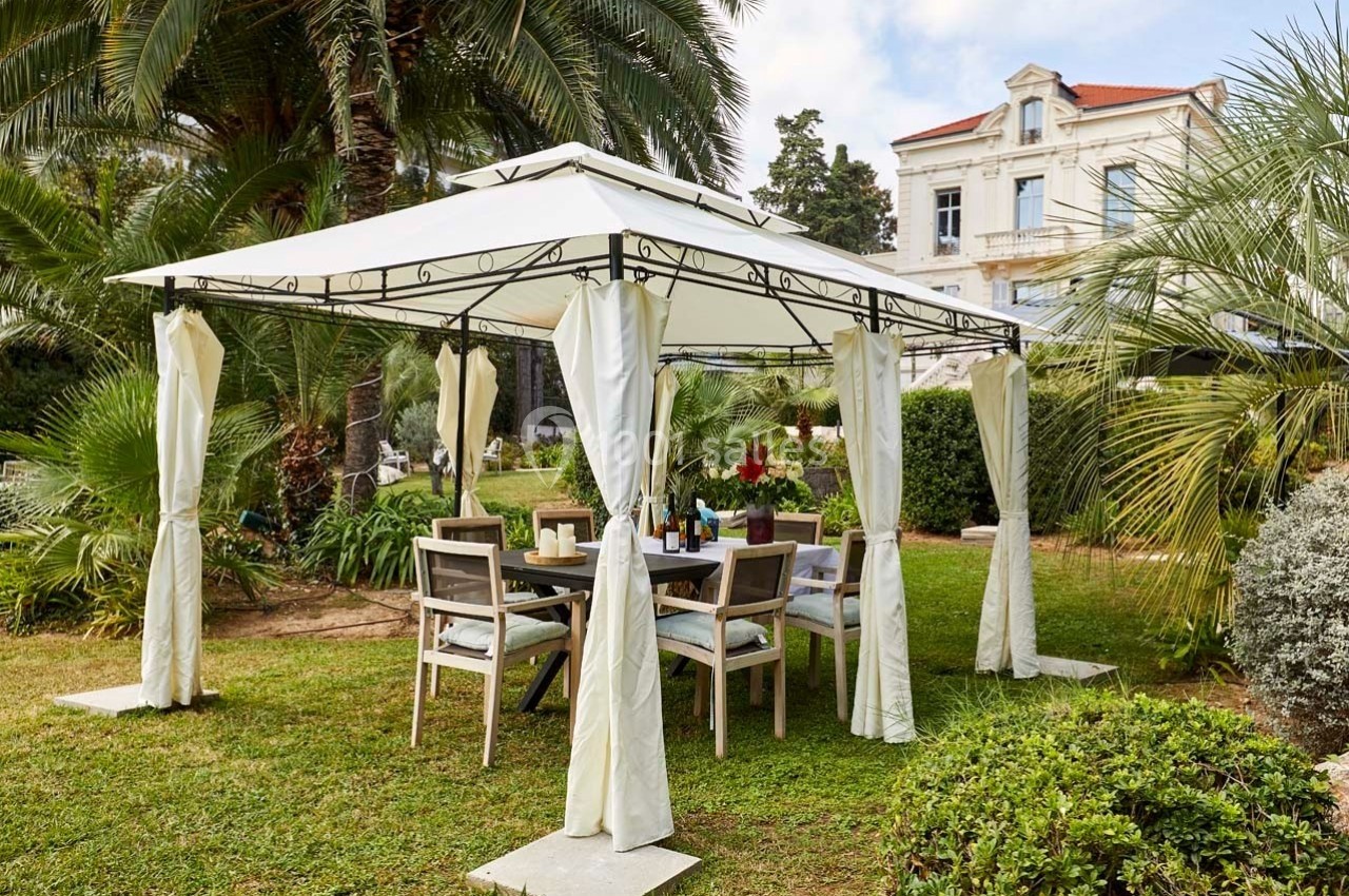 Tonnelle blanche avec rideaux entourée de mobilier de jardin, située dans un espace vert près d'une grande maison.