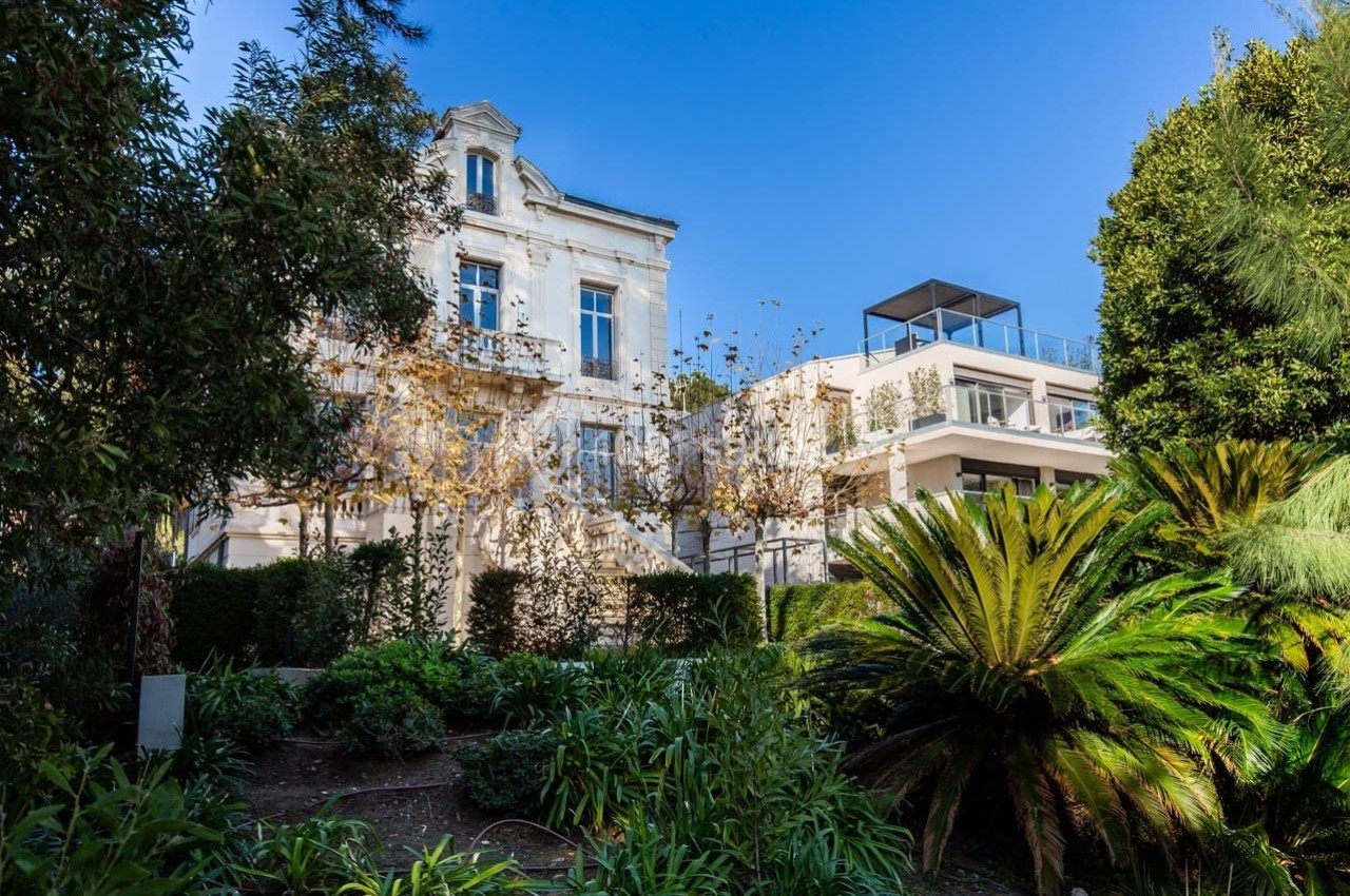 Façade d'une maison ancienne entourée de végétation luxuriante, avec un bâtiment moderne adjacent.