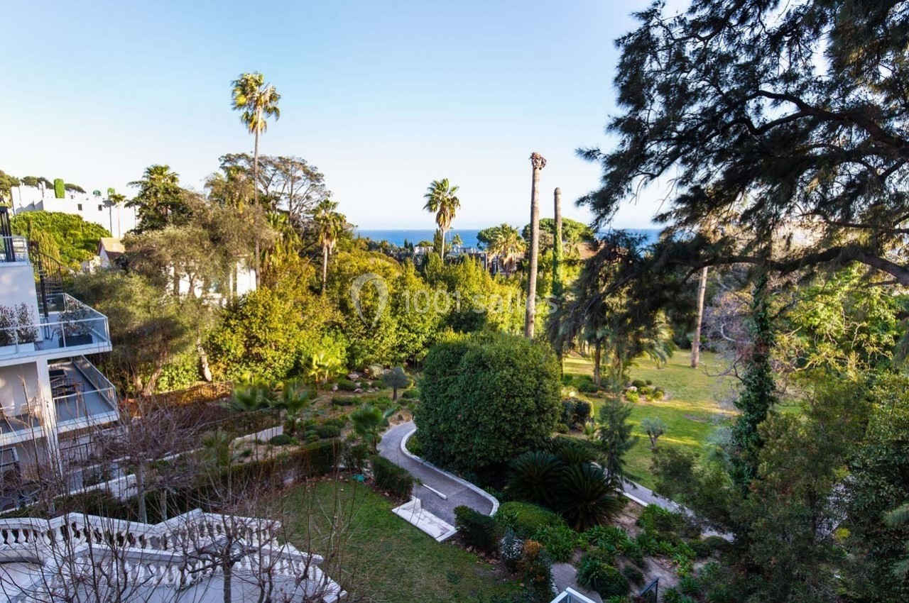 Vue sur un jardin arboré avec des palmiers, des allées et la mer visible à l'horizon sous un ciel dégagé.