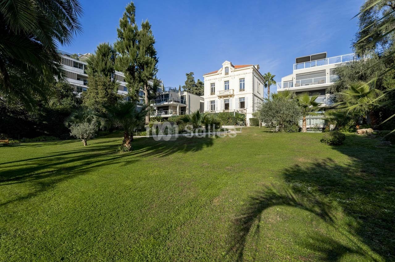 Grande maison blanche entourée d'un vaste jardin verdoyant, avec des arbres et des immeubles modernes en arrière-plan.
