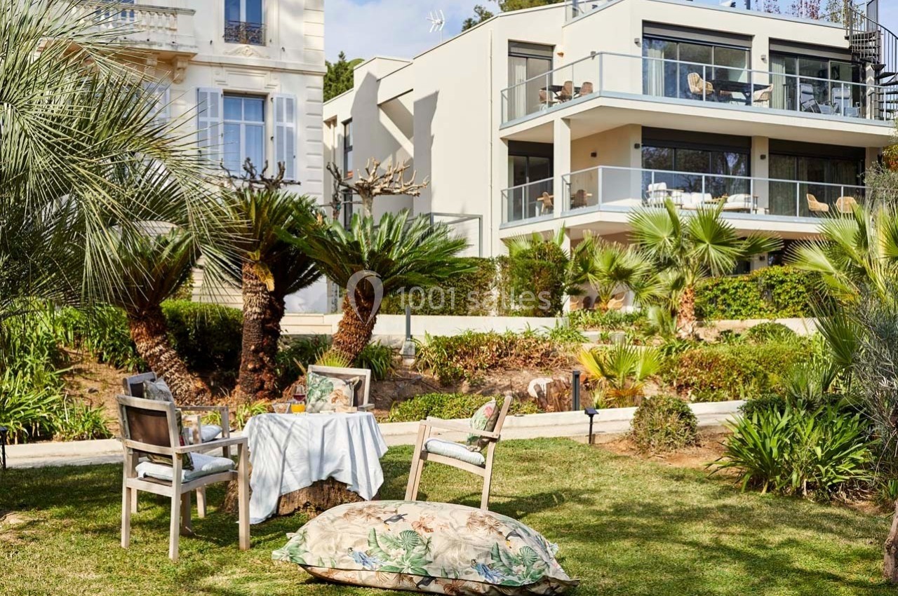 Jardin aménagé avec table et chaises en bois, coussin floral au sol, entouré de palmiers et bâtiments modernes en arrière…