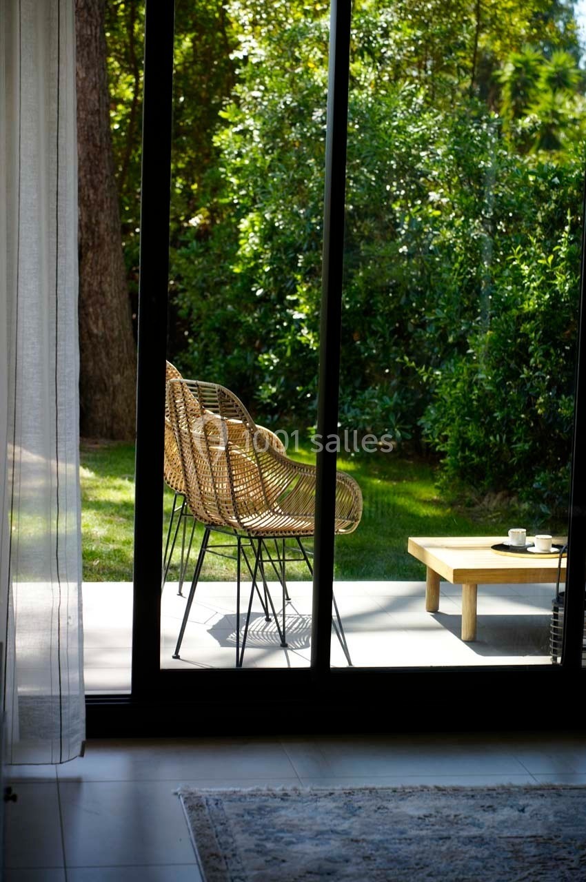 Deux fauteuils en rotin et une table basse avec une tasse, sur une terrasse donnant sur un jardin verdoyant.