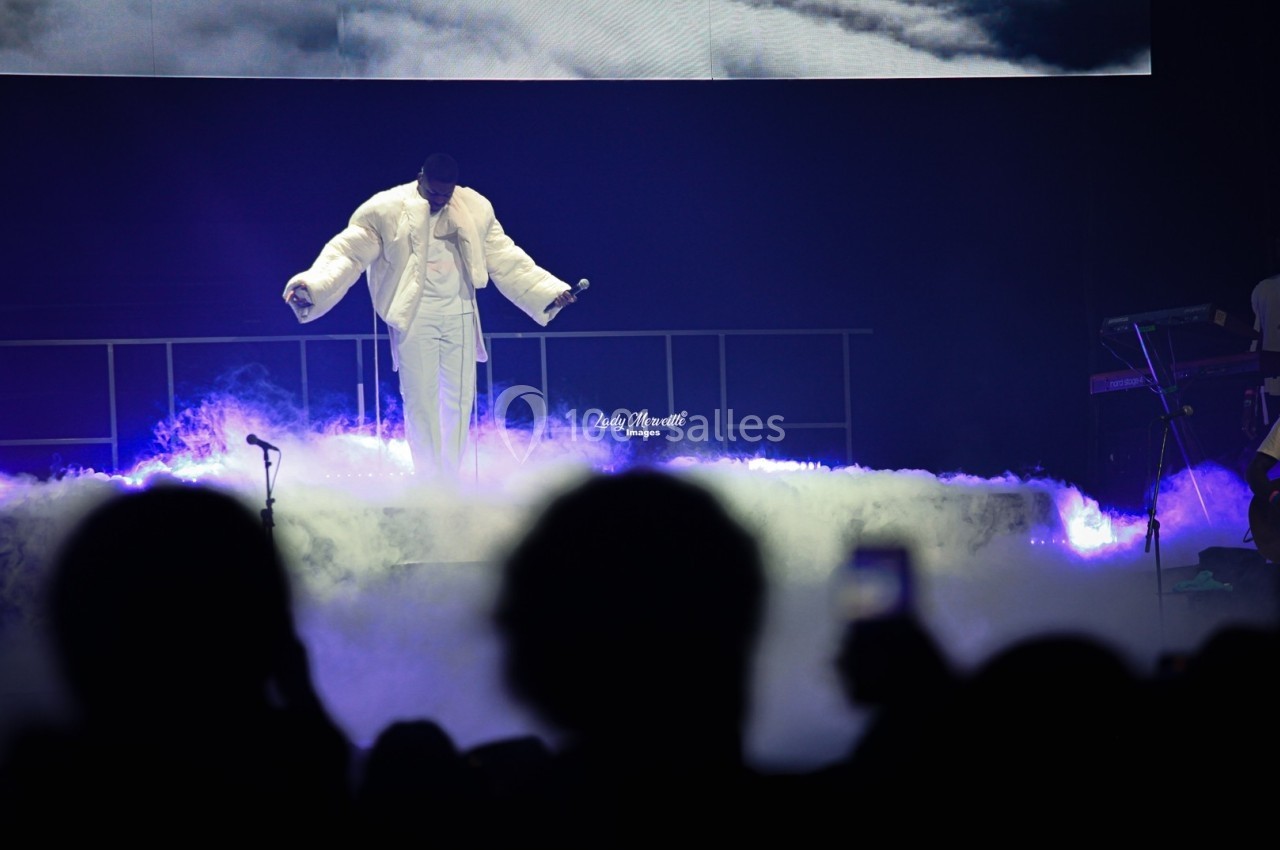 Un artiste en tenue blanche se produit sur scène entouré de fumée, avec un public en contre-jour au premier plan.