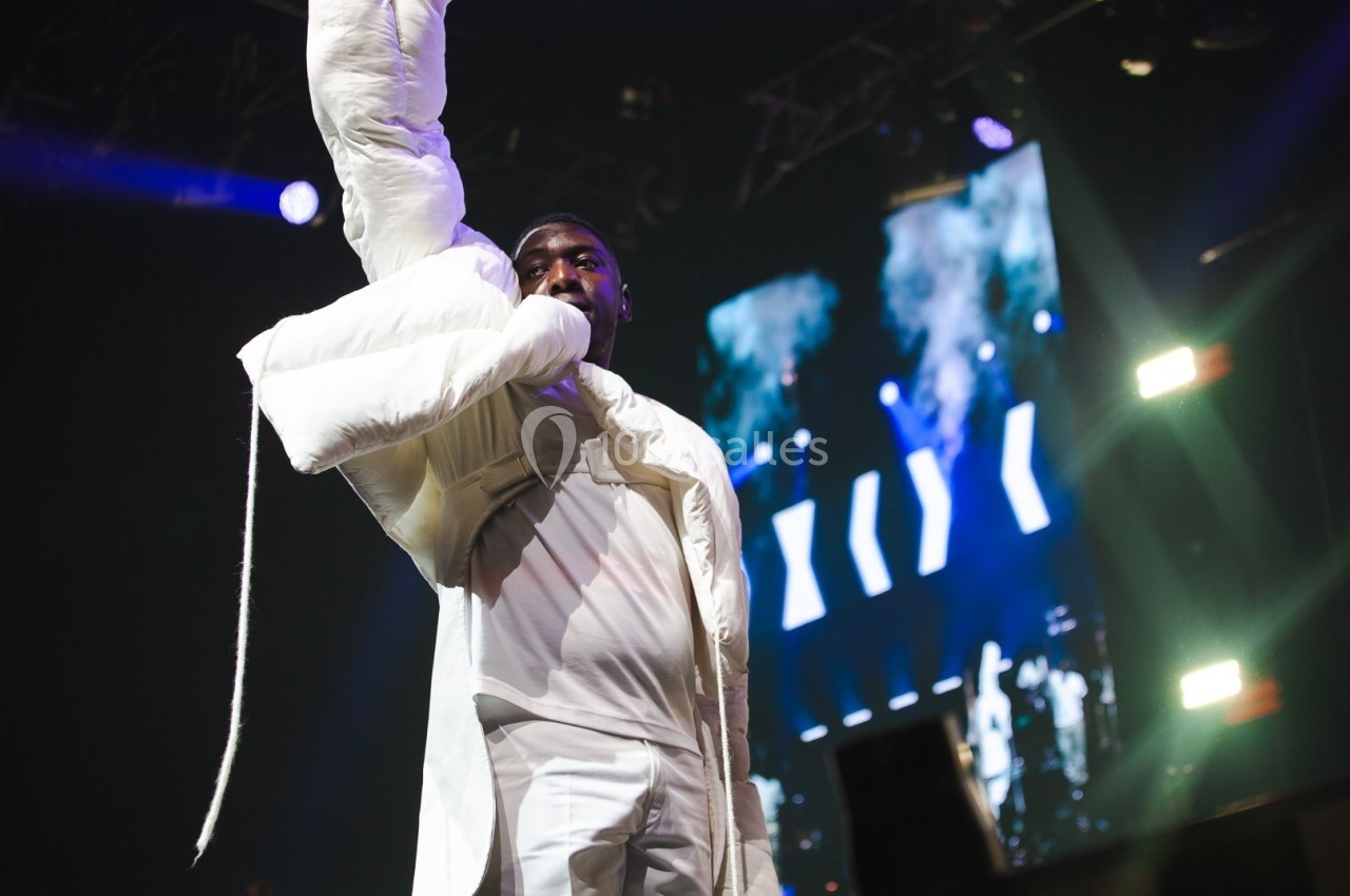 Un artiste en tenue blanche performe sur scène sous des éclairages vifs, avec un écran lumineux en arrière-plan.