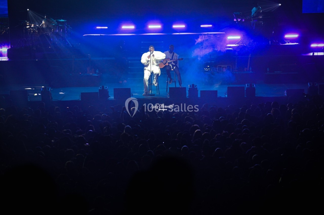 Un chanteur sur scène, éclairé par des projecteurs bleus, devant une foule dans une salle de concert.