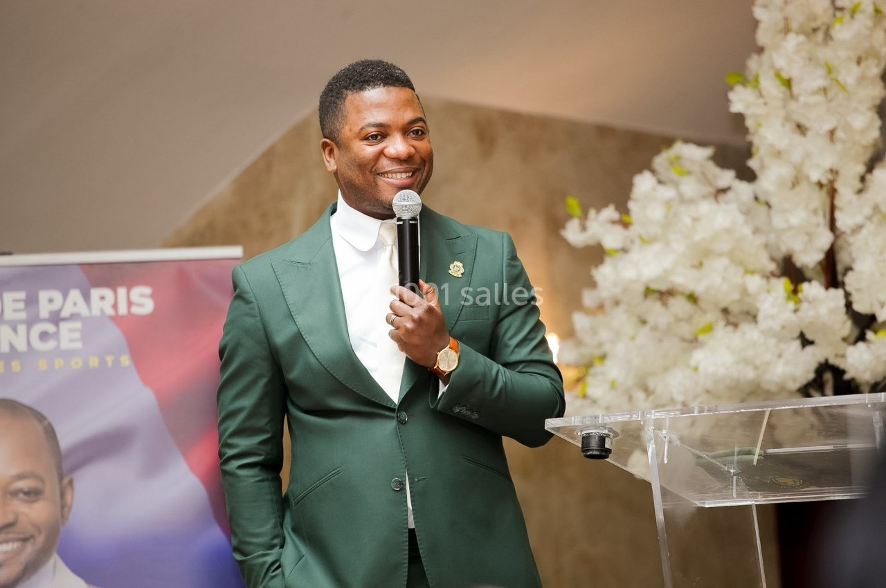 Un homme en costume vert parle au micro lors d'un événement, debout près d'un pupitre et d'une décoration florale blanche.