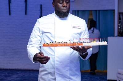 Un chanteur sur scène, vêtu de blanc, levant un bras sous des lumières de spectacle et un écran en arrière-plan.