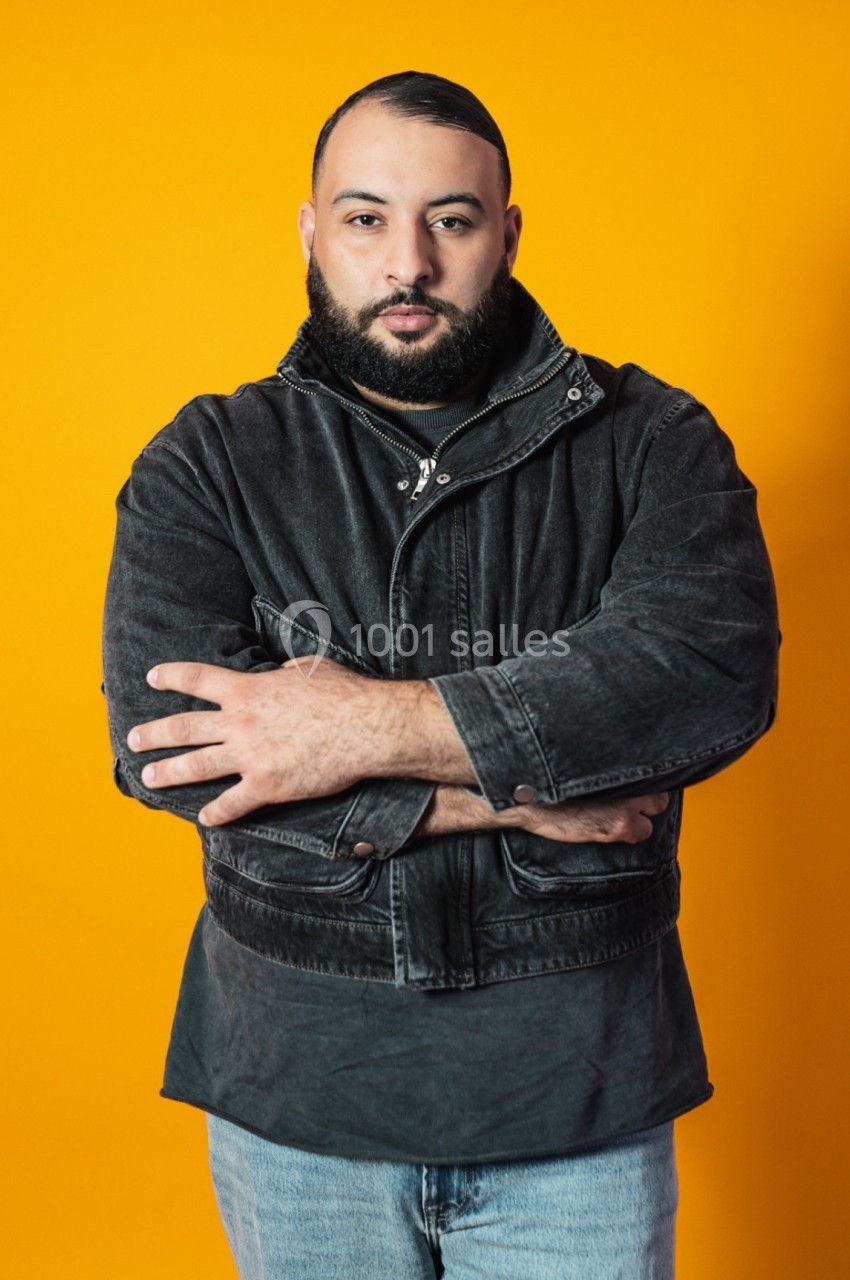 Homme debout, bras croisés, portant une veste en jean foncé, sur un fond orange uni.