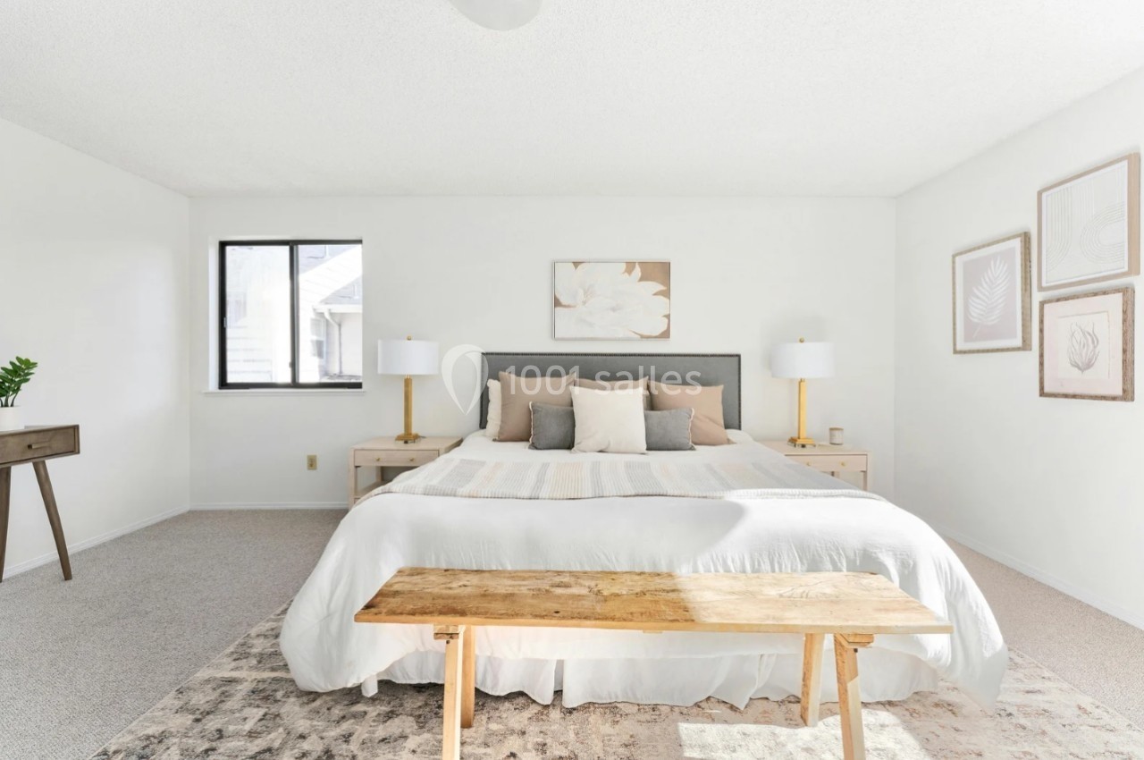 Chambre lumineuse avec lit double, tête de lit grise, tables de chevet, lampes dorées et banc en bois au premier plan.