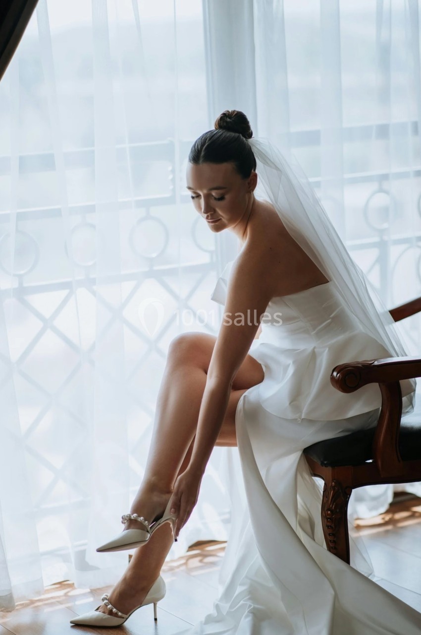 Une mariée assise ajuste ses chaussures, vêtue d'une robe blanche avec un voile, près d'une fenêtre lumineuse.